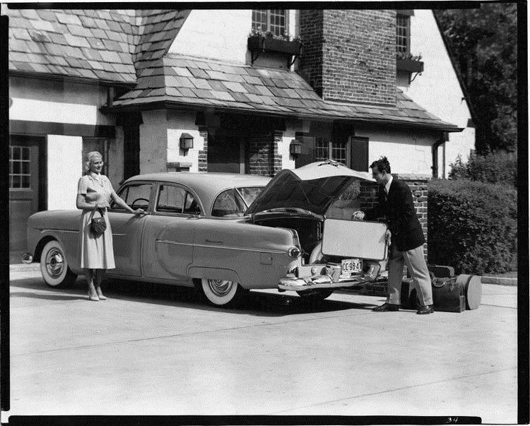 1951 Packard sedan, female standing at driver's door, man loading luggage into trunk